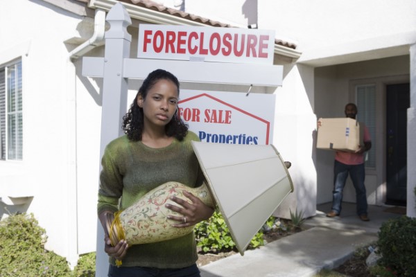 Many 'We Buy Houses' scams cost homeowners substantial amounts of lost equity. Distressed woman standing next to a 'Foreclosure' sign in her front yard.